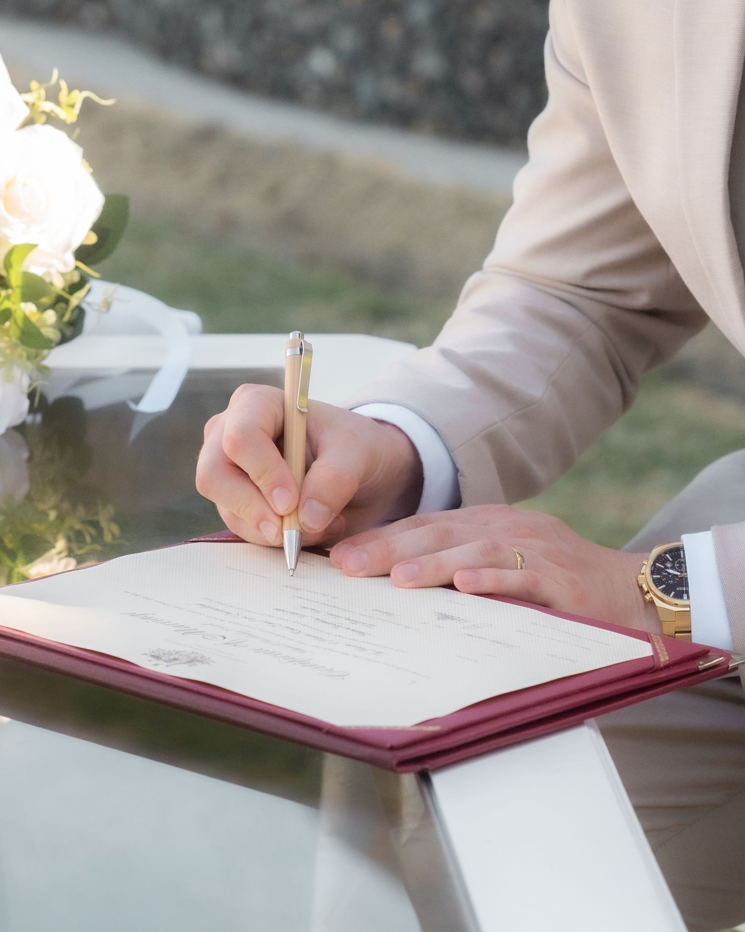 Groom in a beige suit signs the marriage certificate with a gold pen beside a white floral bouquet.