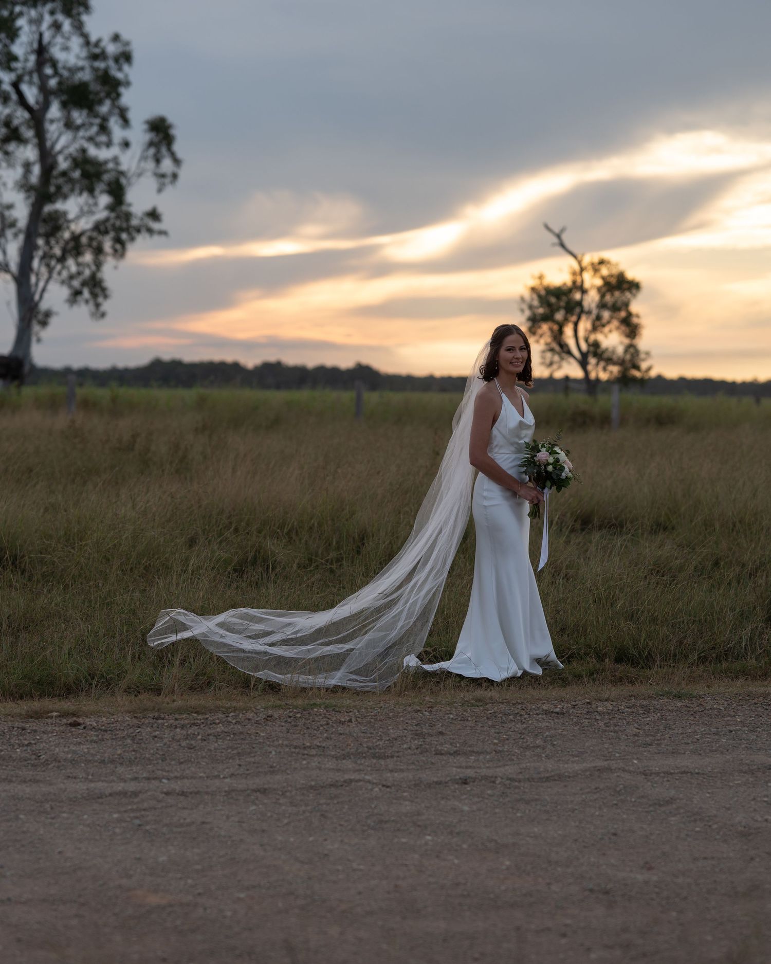Bride in a white gown with a long flowing veil holds a bouquet on a country road at sunset.