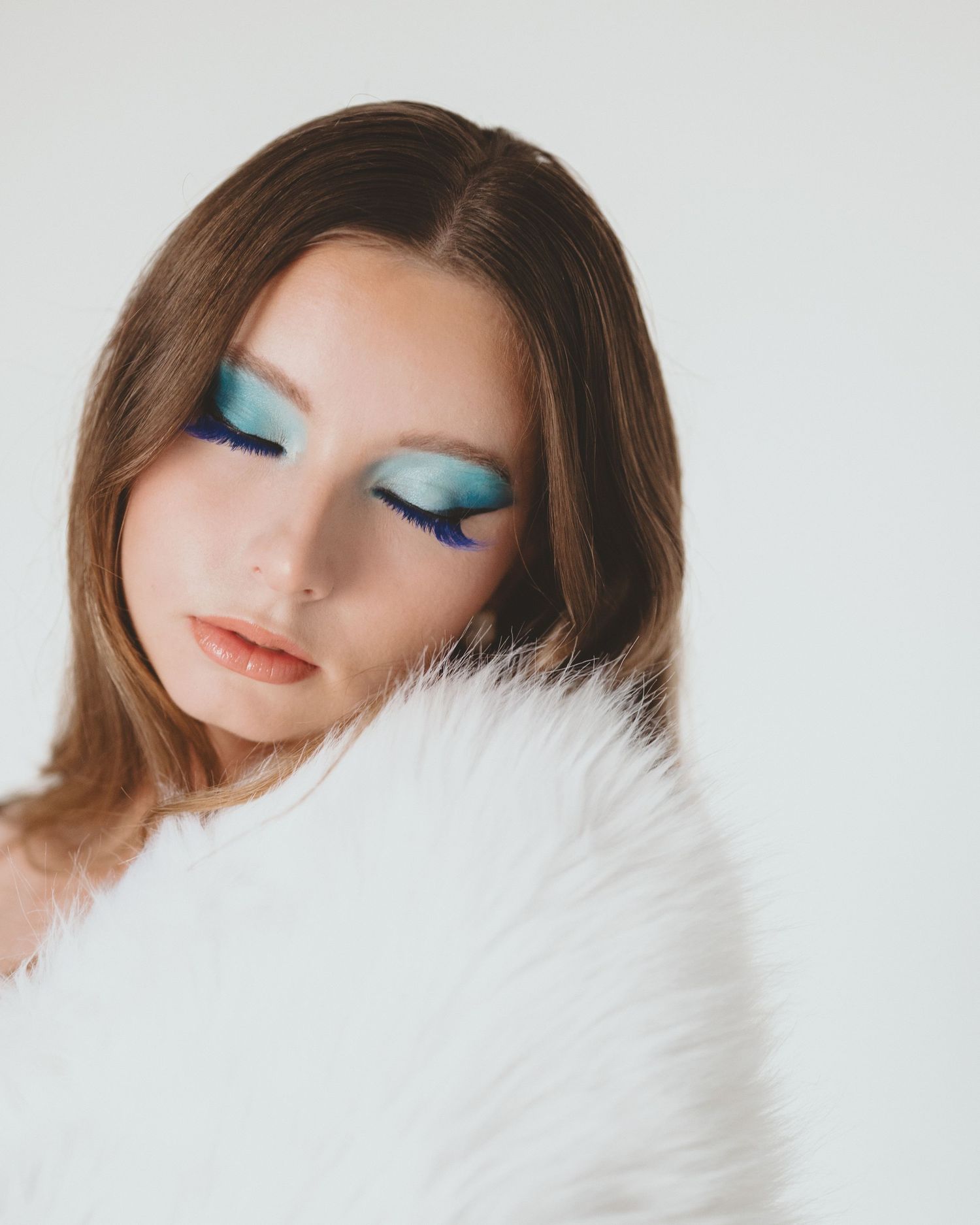 Woman with eyes closed showing teal and blue gradient eyeshadow and dramatic lashes, resting her chin on a white faux fur stole.