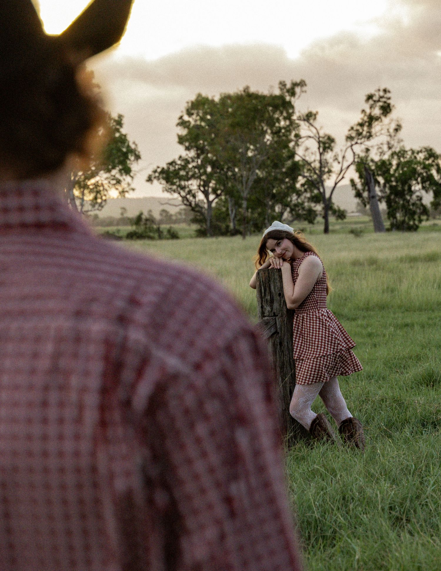 Woman in a gingham dress and white head scarf leans on a wooden fence post in a grassy field as a cowboy watches from the foreground.