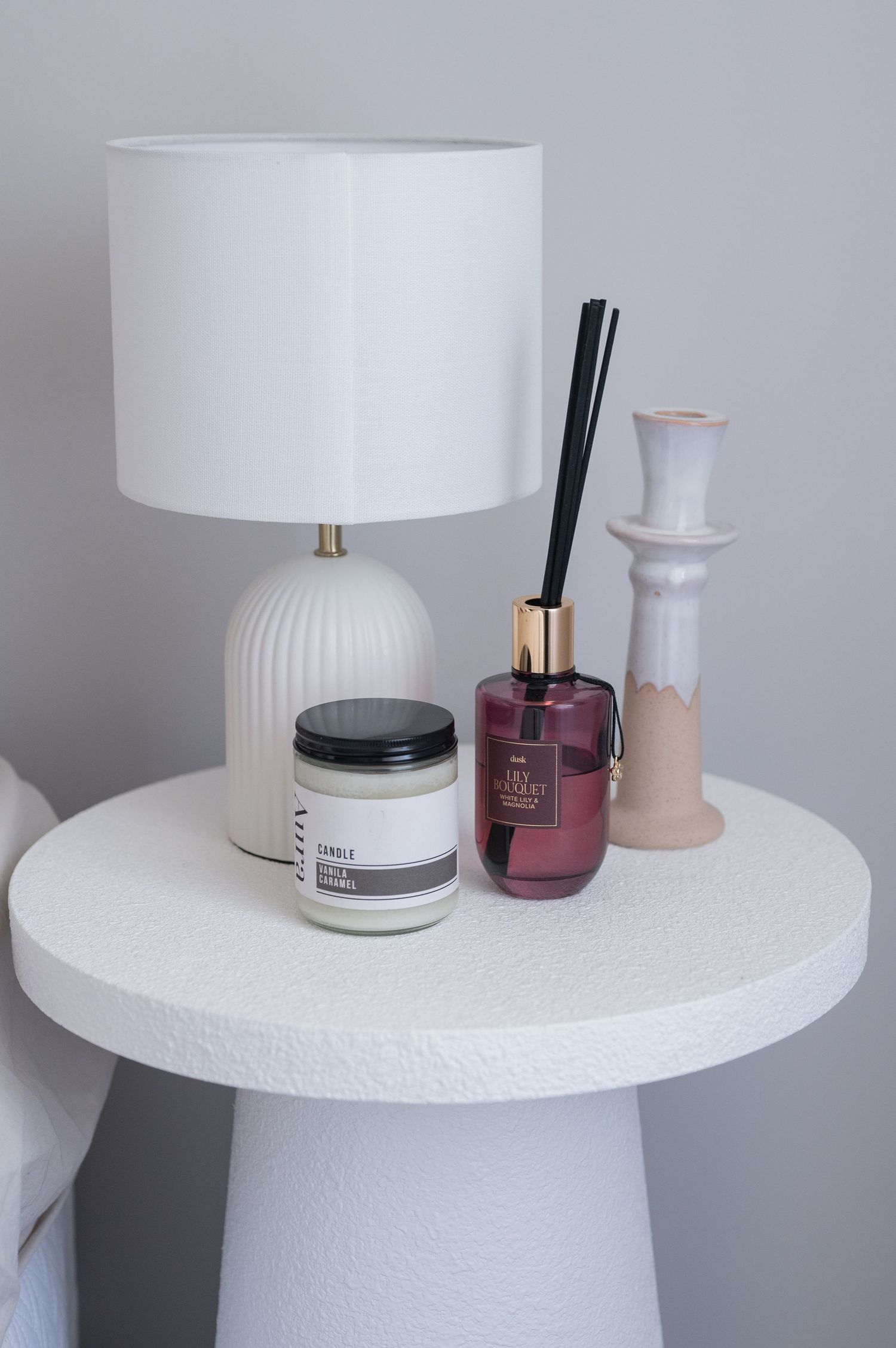 White bedside table styled with a ribbed lamp, vanilla caramel candle, reed diffuser and ceramic candle holder.