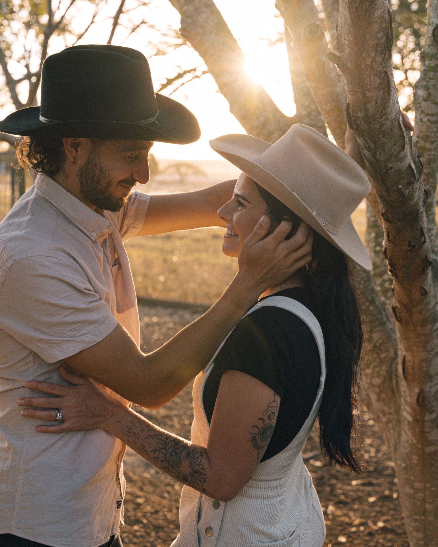 Couple in cowboy hats share a tender moment under a tree as golden sunlight filters through the branches.