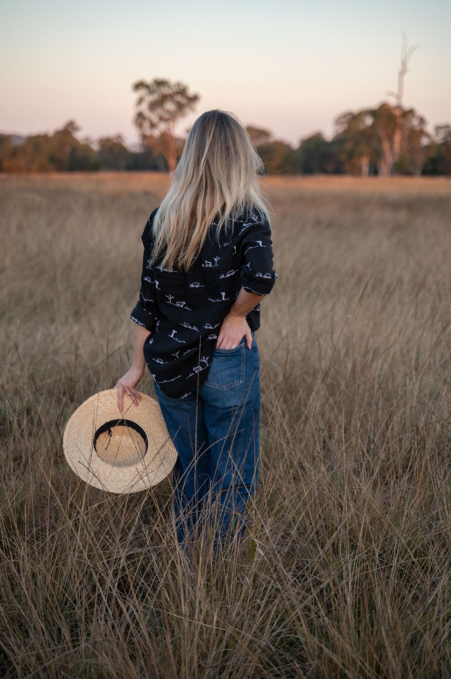 A woman with long blonde hair stands in a golden field at sunset, holding a straw hat and wearing a navy printed linen shirt.