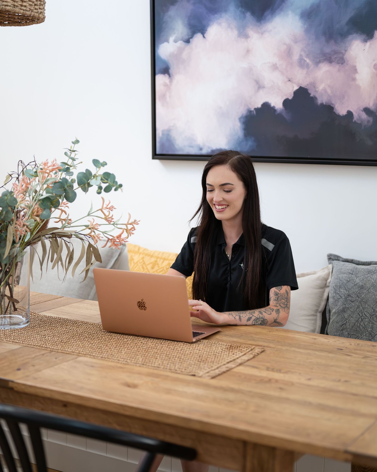 Smiling woman with tattooed arm working on a rose gold laptop at a rustic wooden table beside a floral arrangement.