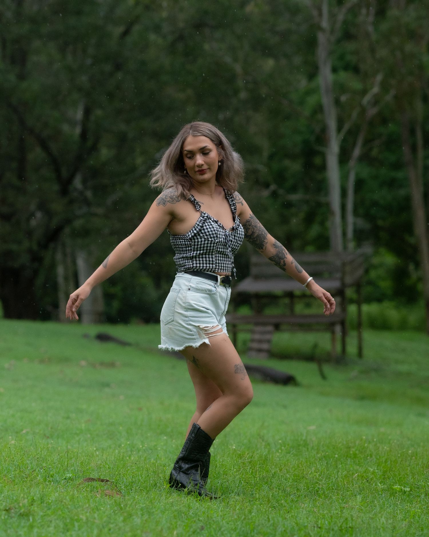 Woman in a gingham top, denim shorts and black boots dancing on a green lawn with a wooden bench and trees behind her.