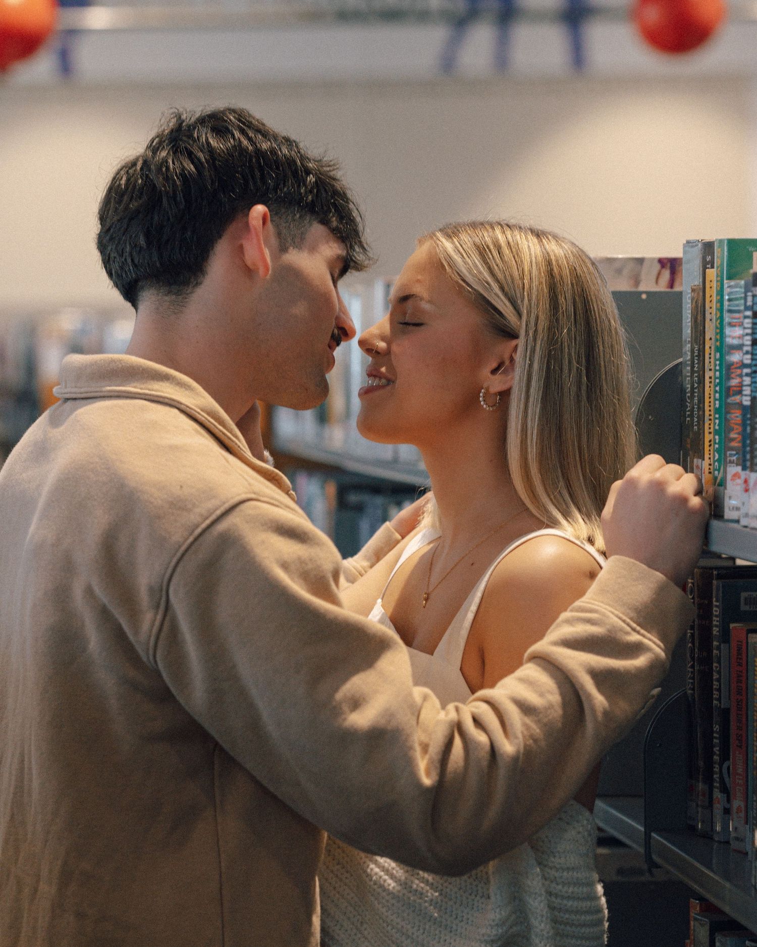 A couple smiles and leans in close beside a library bookshelf in a warm, romantic moment.