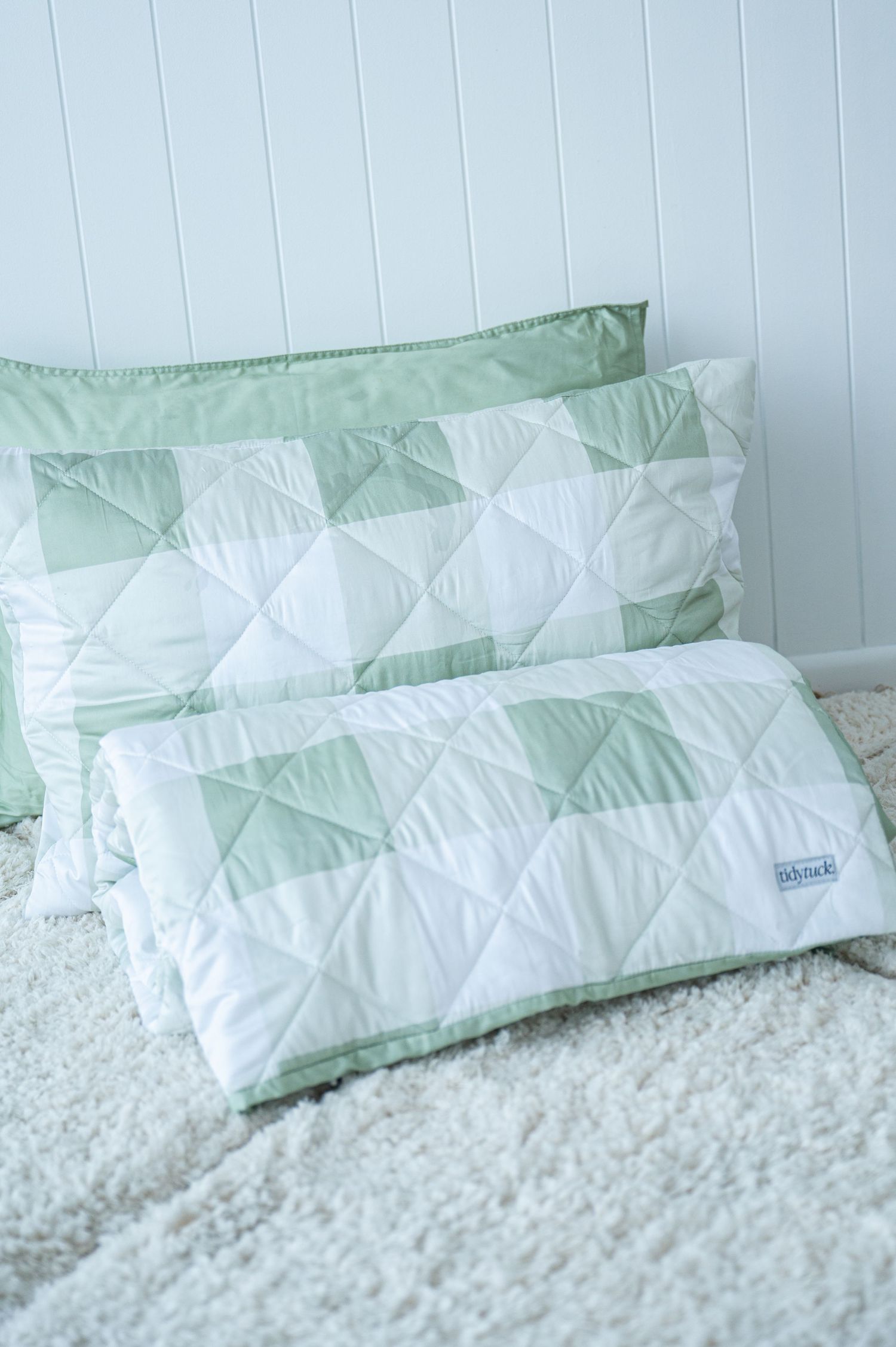 Sage green and white checkered quilt rolled up in front of matching pillows on a fluffy cream rug against white paneled walls.