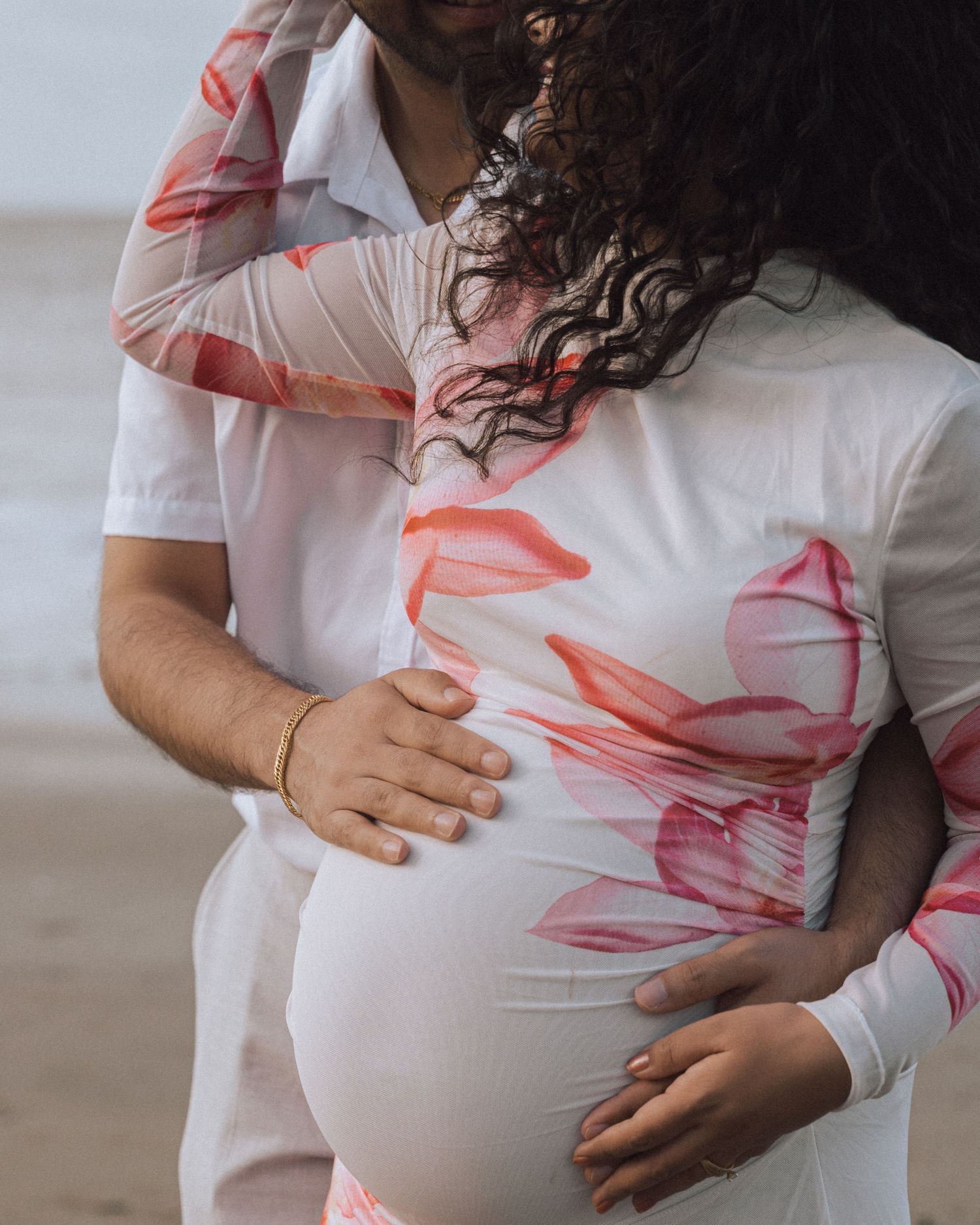 Close-up of a couple's hands resting on a pregnant belly in a white floral maternity dress on the beach.