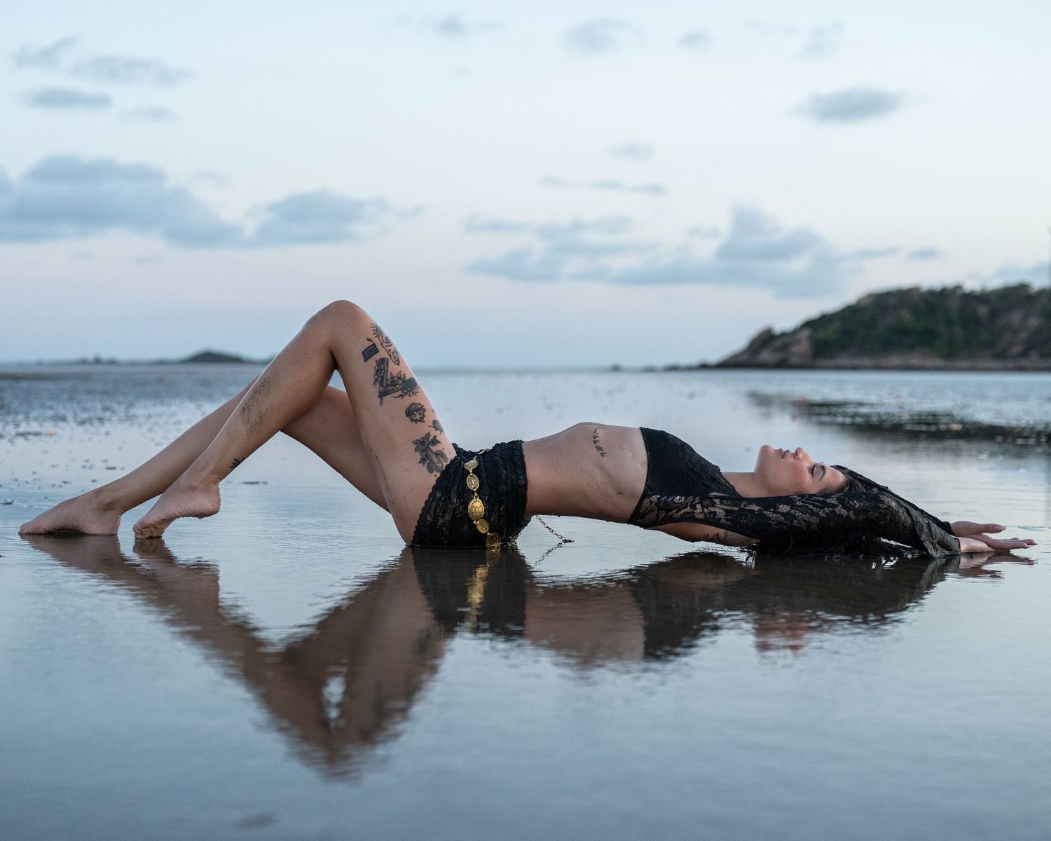 Tattooed woman in black lace lying on a reflective wet beach at sunset, her reflection mirrored in the shallow water.