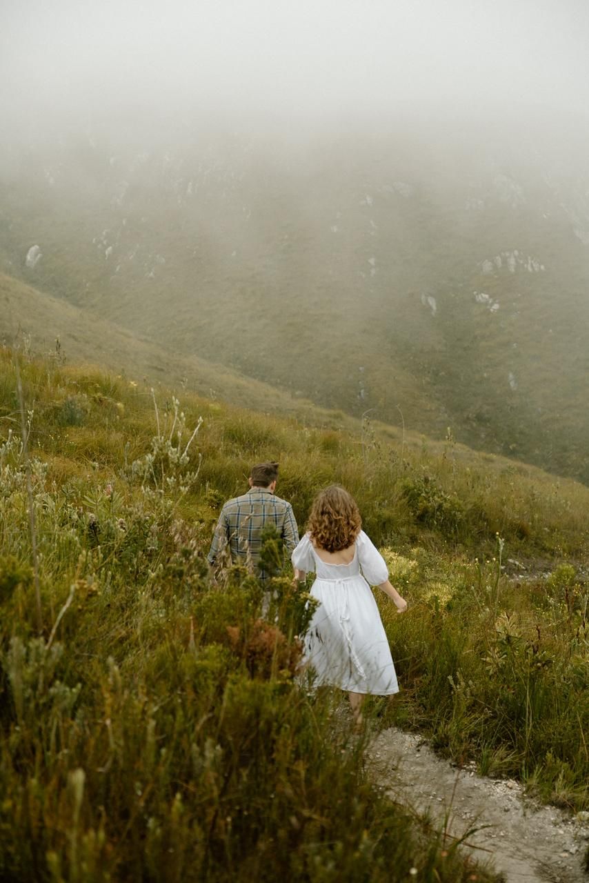 Couple walking up a misty mountain trail through lush green grass in foggy conditions.