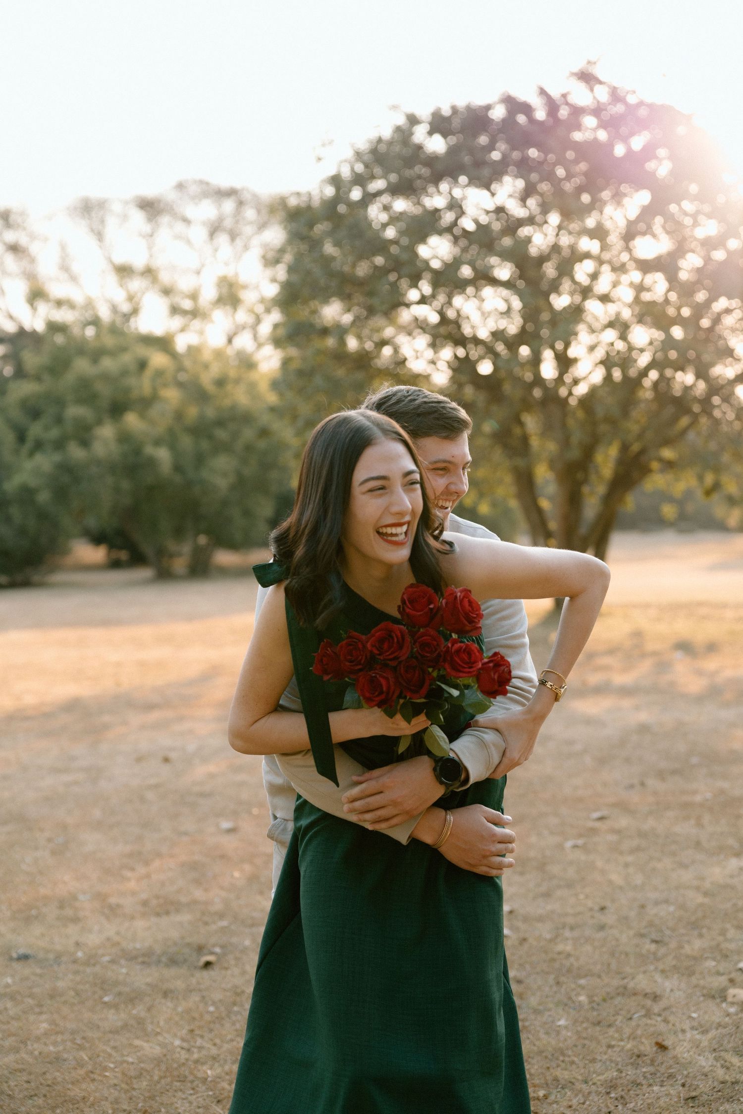 Joyful woman in green dress laughing while holding red roses, embraced by partner in sunny park setting.