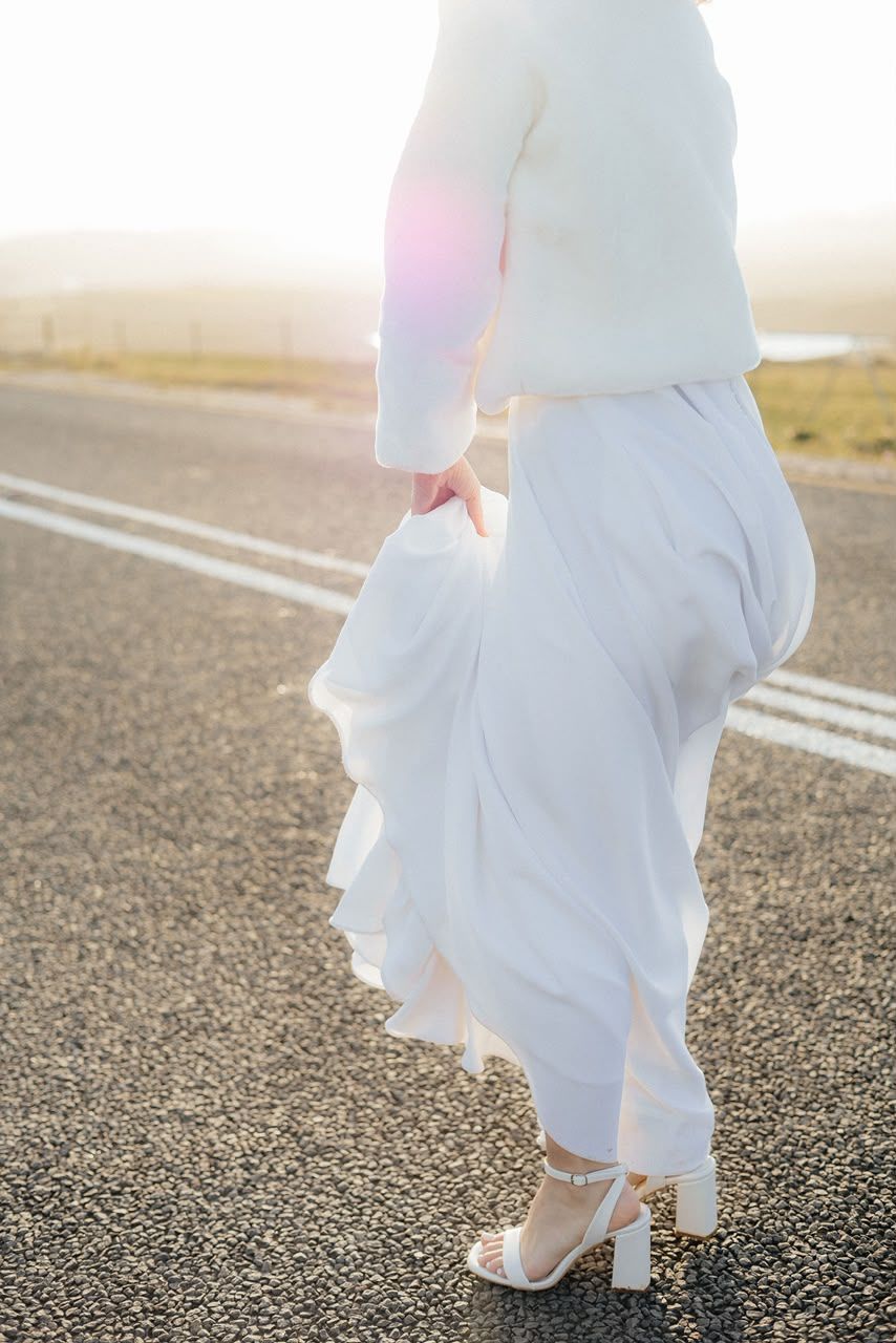 Bride in white ruffled skirt, cropped jacket, and block heel sandals walking on a sunlit road.