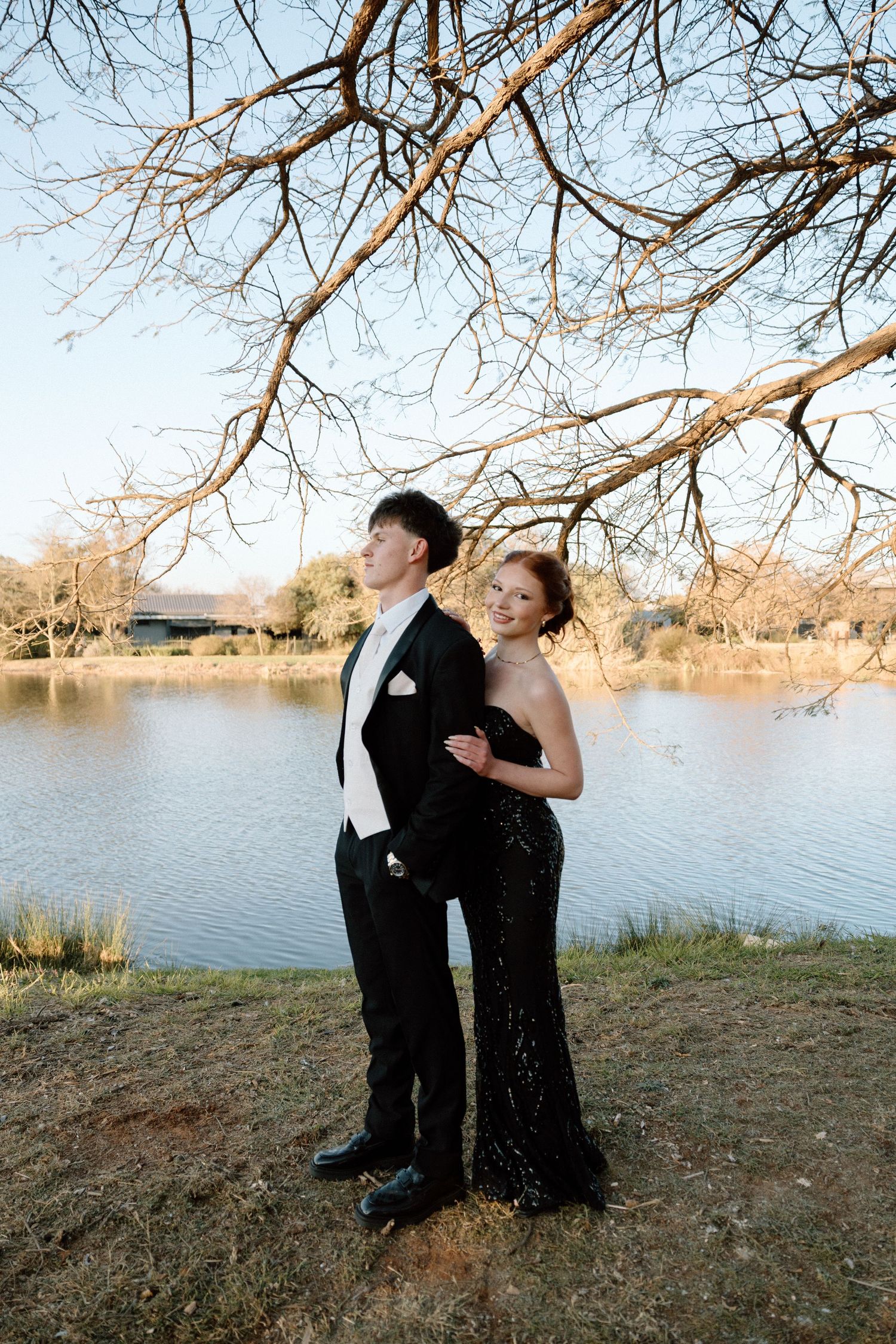 Elegant couple in black formal wear posing by a serene lake under bare winter tree branches.