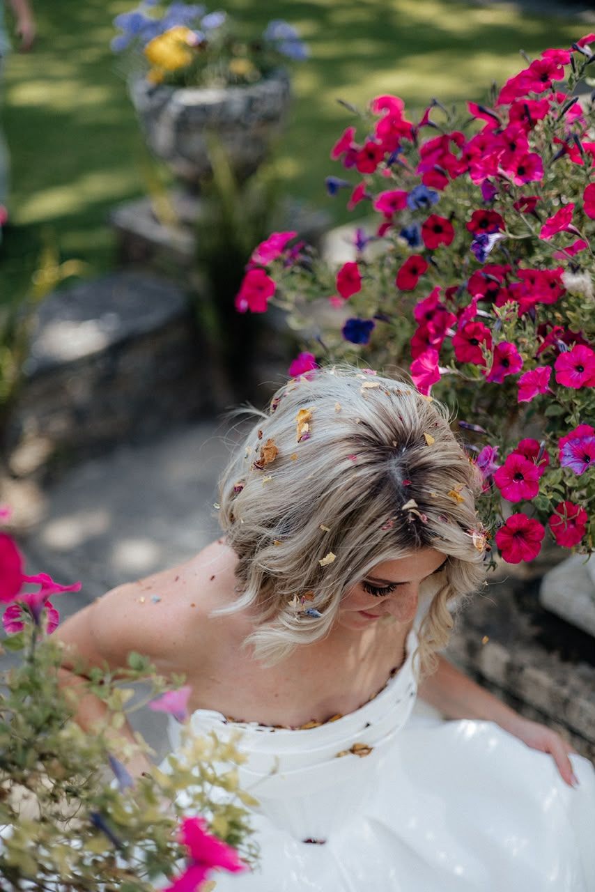 Bride in white dress surrounded by vibrant pink petunias in a garden, with confetti in her updo hair.