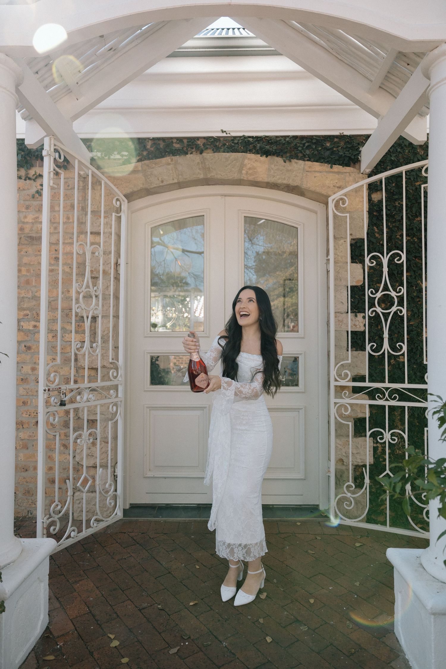 Woman in white lace dress holding champagne bottle, laughing at elegant white arched doorway with ornate iron gates.