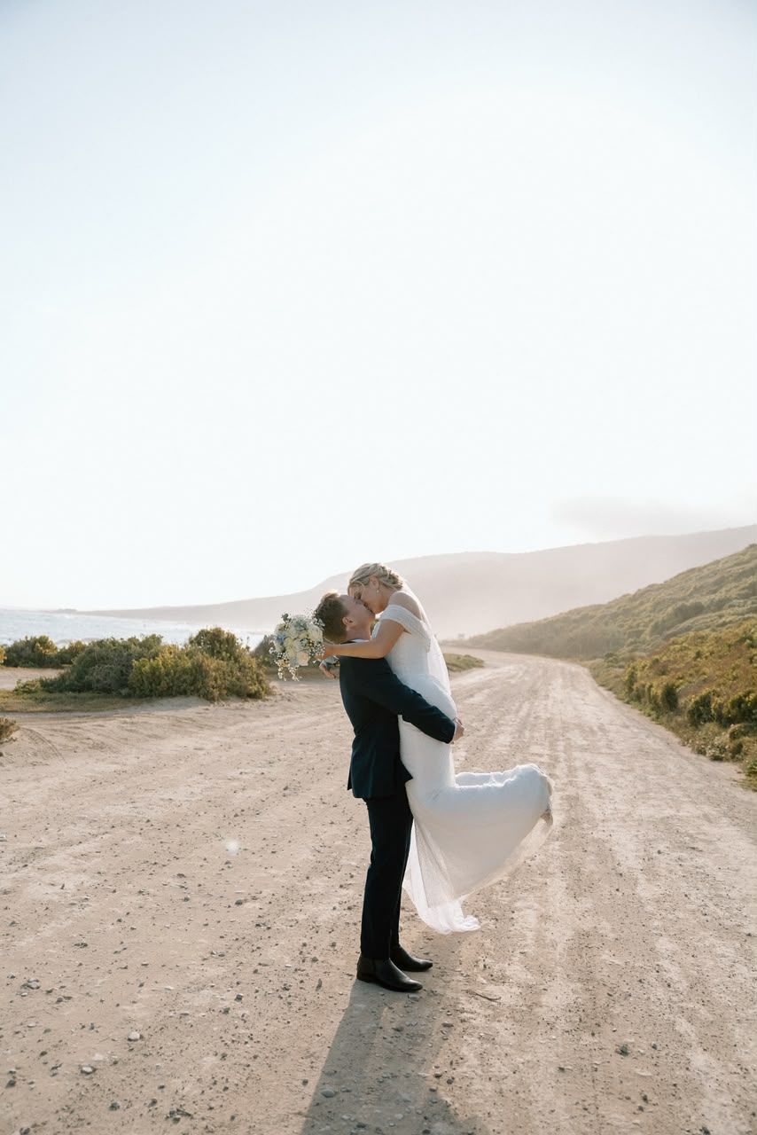 Groom lifting and kissing bride in flowing white dress on scenic coastal dirt road with bright sky backdrop.