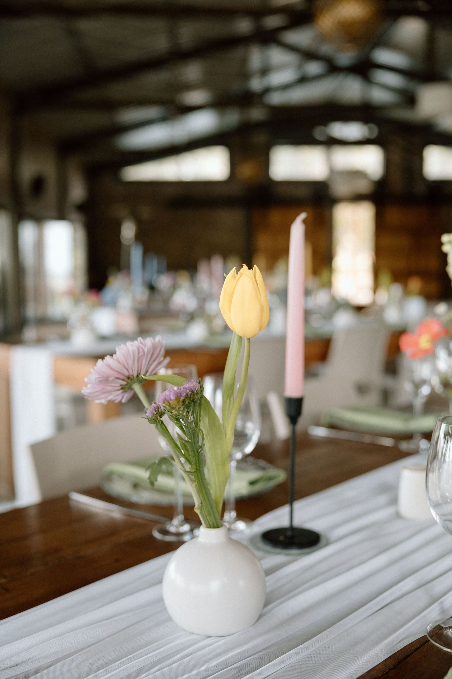 White bud vase with yellow tulip and pink flowers beside a candle on a wooden wedding reception table.