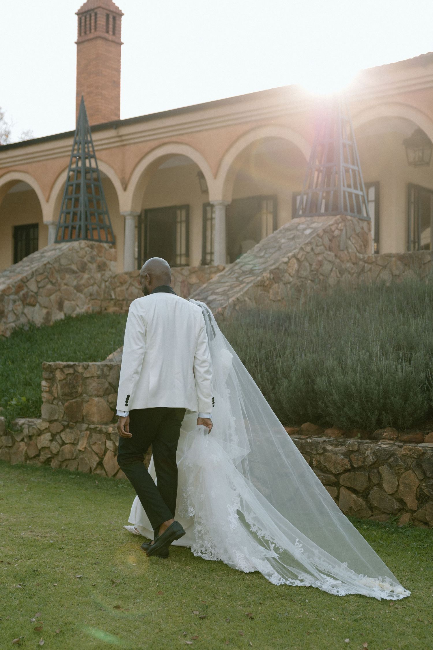 Bride and groom walking toward elegant stone villa with arched colonnade, wedding veil trailing gracefully on green lawn.