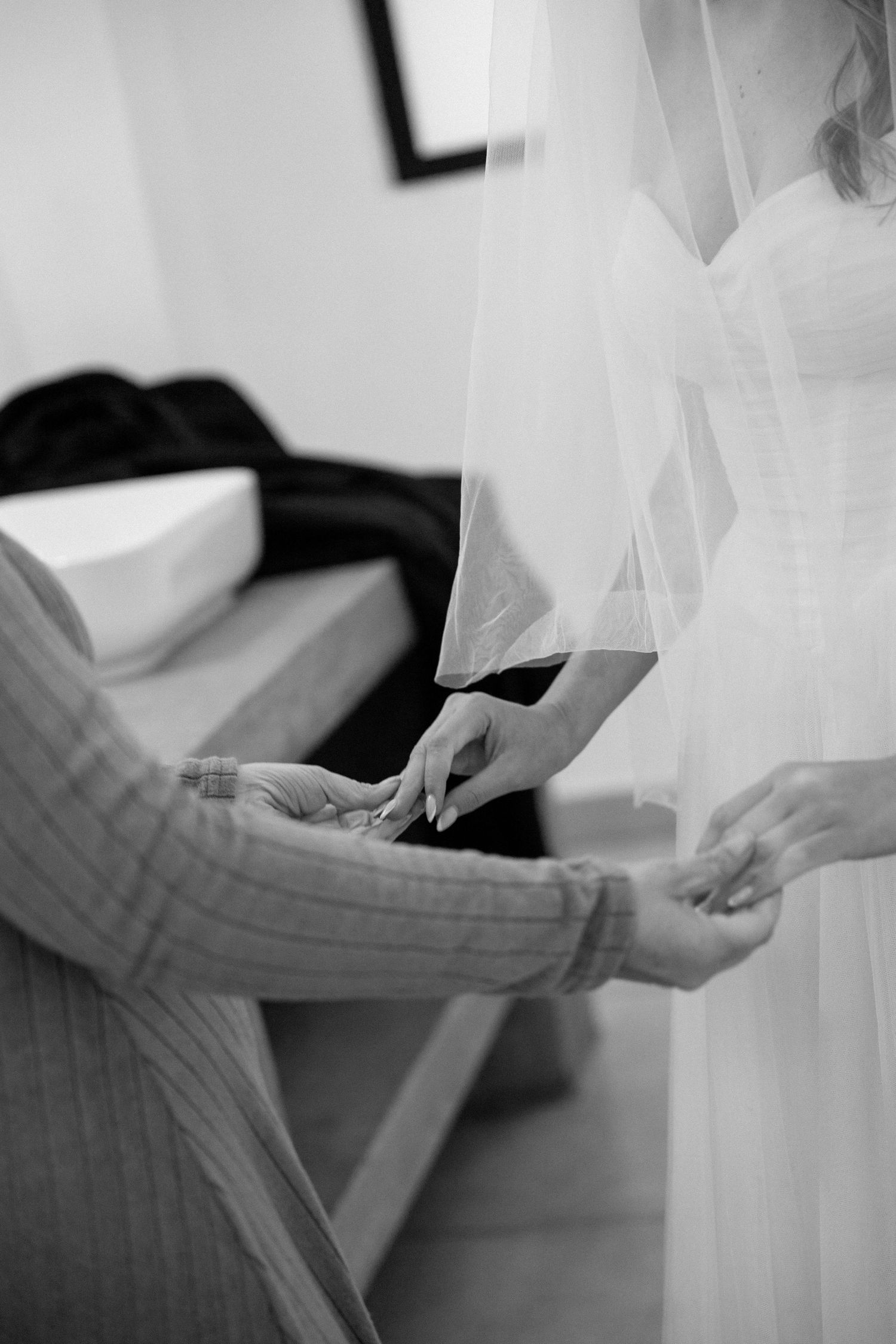 Black and white image of bride in veil having dress fastened by helper in bridal suite.