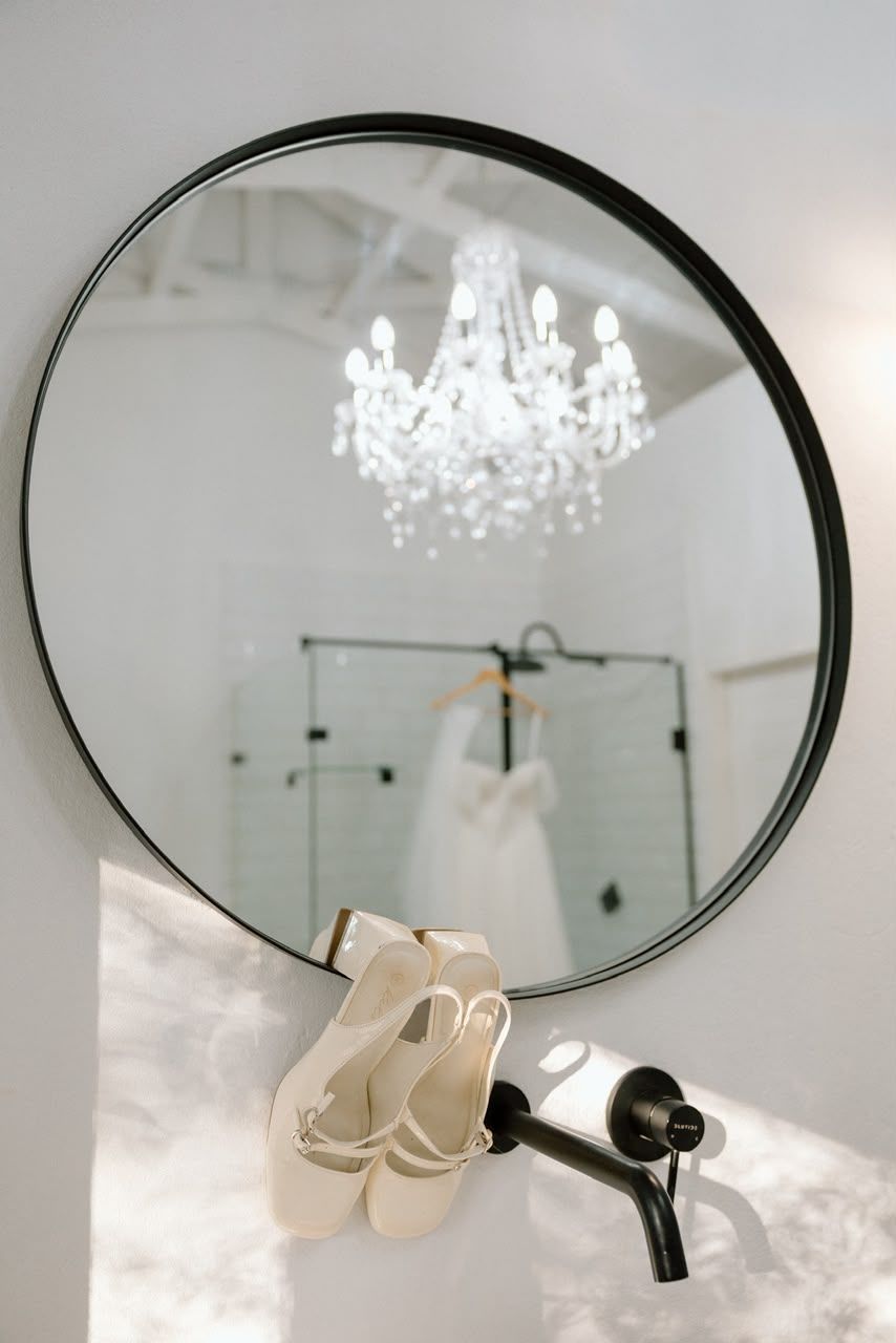 Elegant bathroom vanity with round black-framed mirror reflecting a crystal chandelier and wedding dress hanging in background.