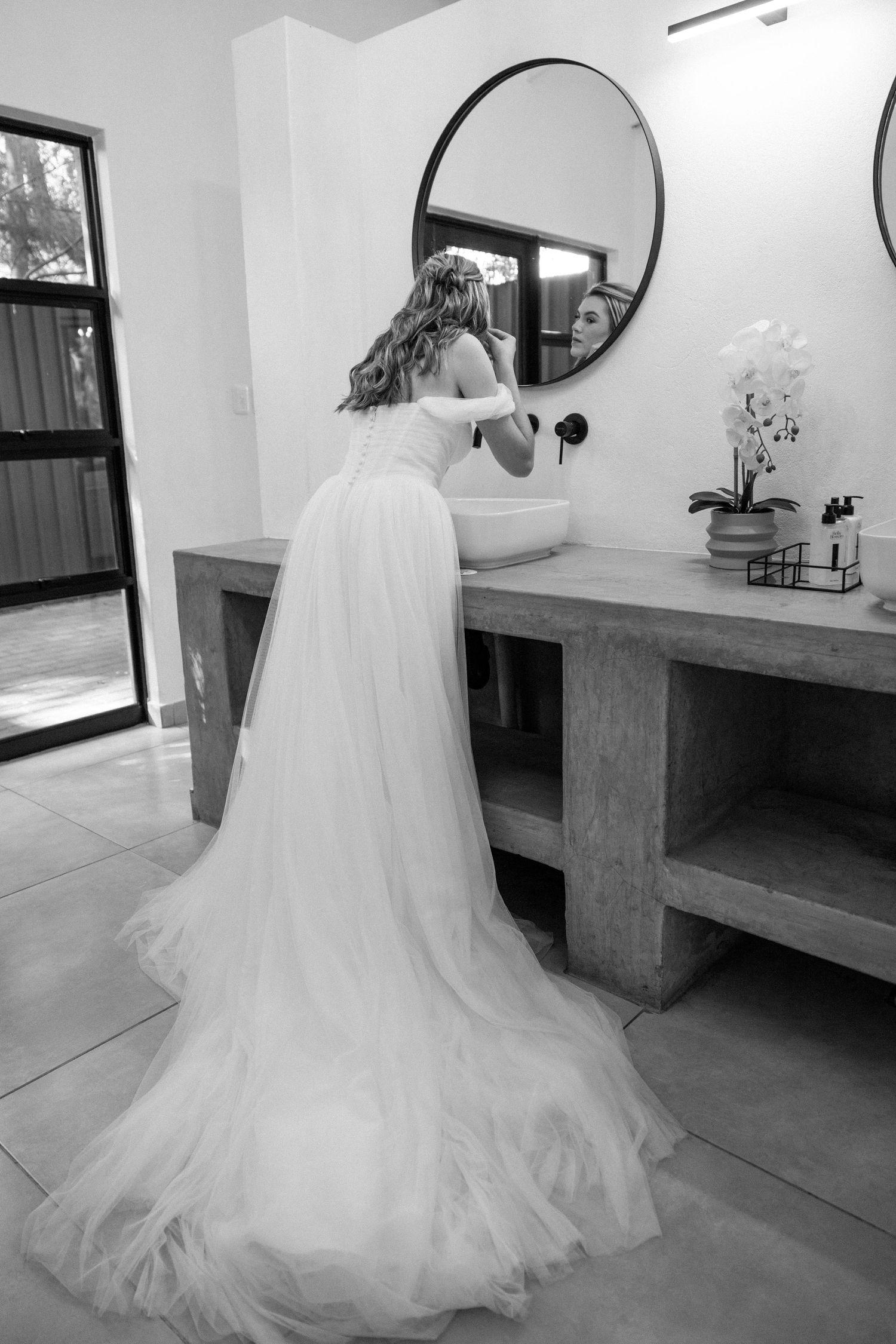 Bride in a flowing off-shoulder tulle gown stands before a round mirror in a modern bathroom.