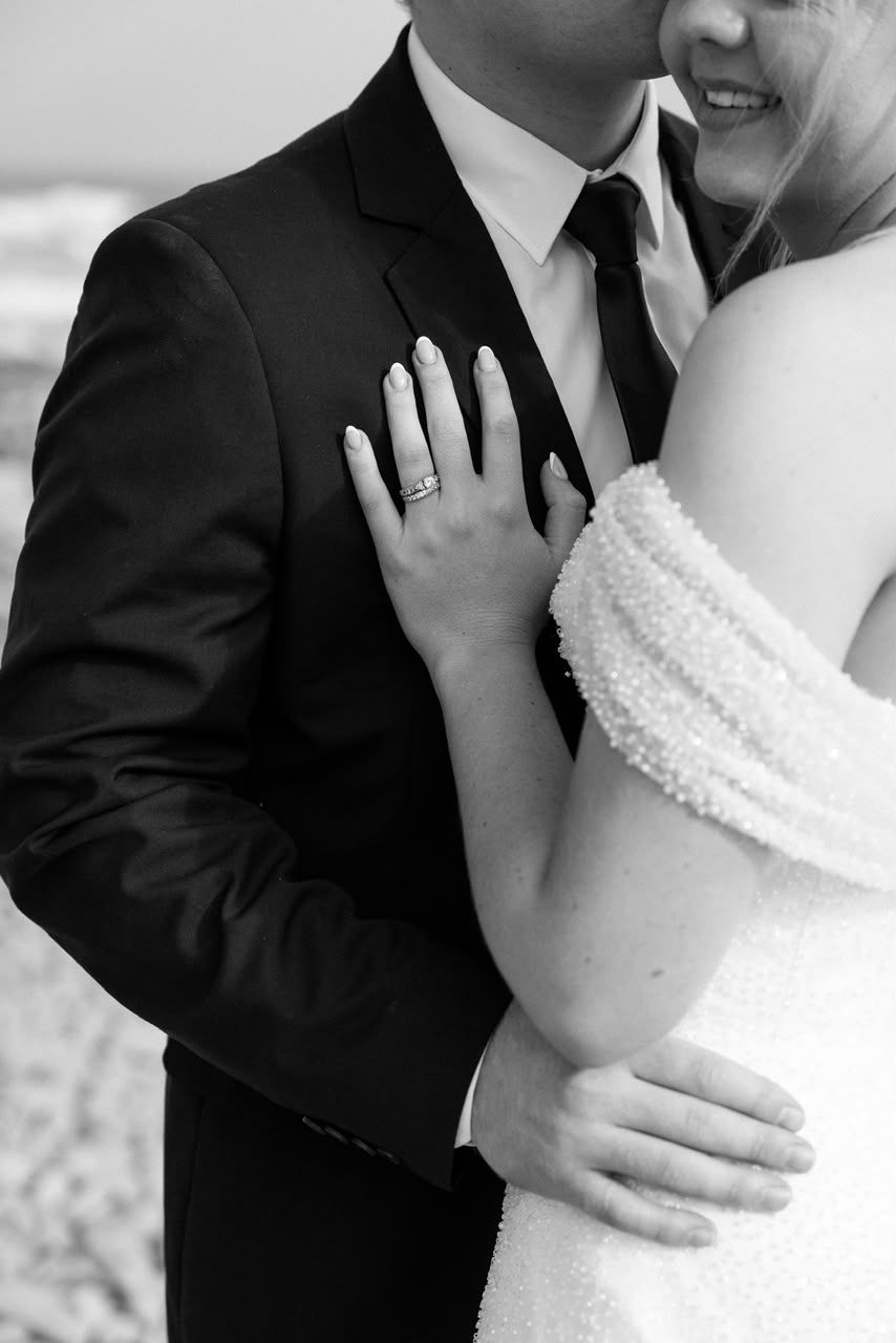 Bride in pearl strapless gown resting hand on groom's black suit, showing diamond ring, in black and white.