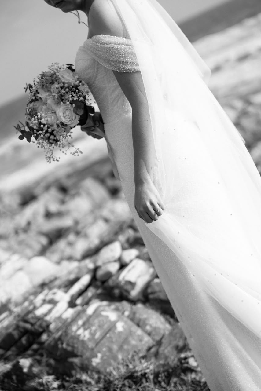 Bride in off-shoulder pearl wedding gown holding floral bouquet on rocky coastal landscape in black and white.