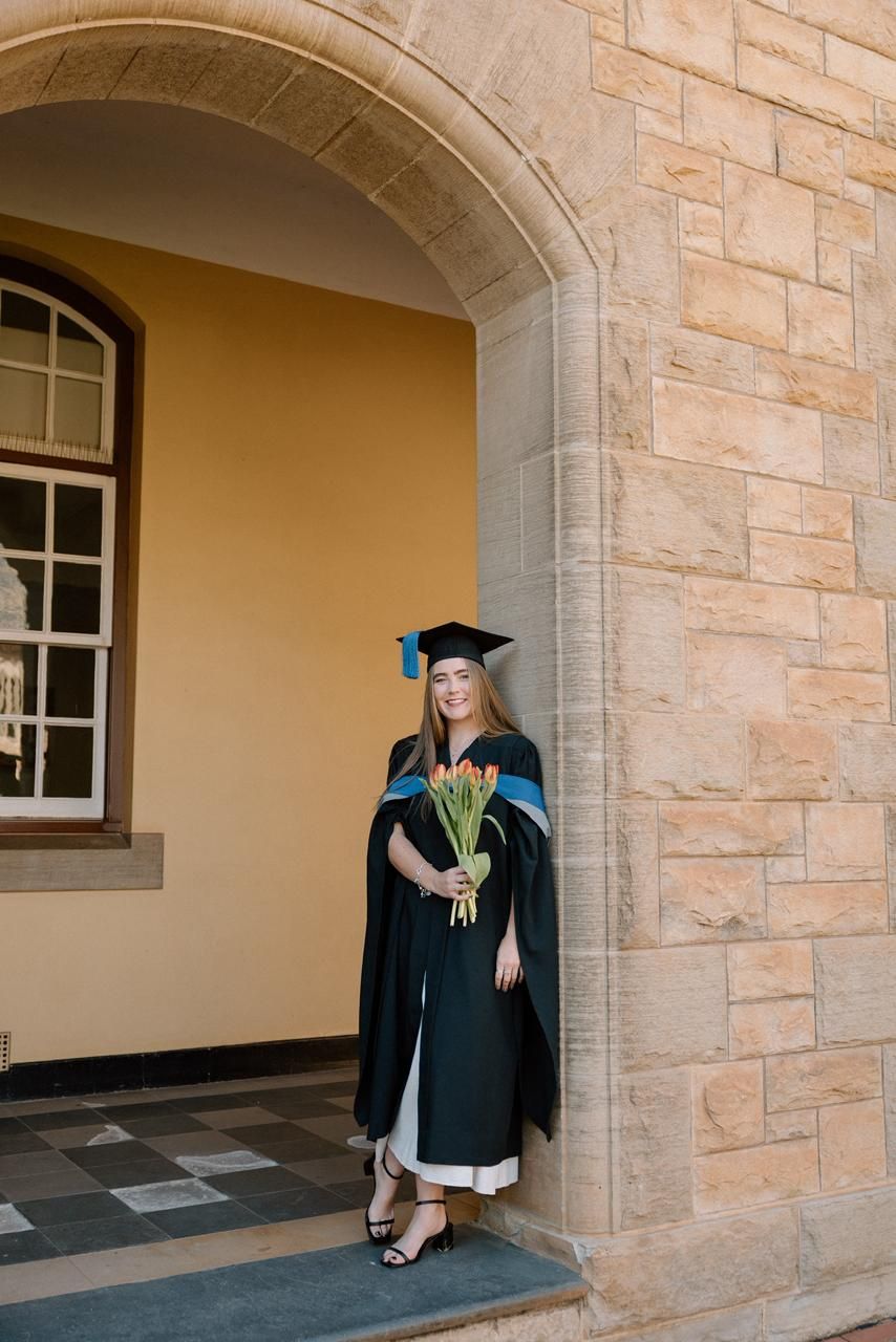 Female graduate in black gown holds tulip bouquet while posing under stone university archway.