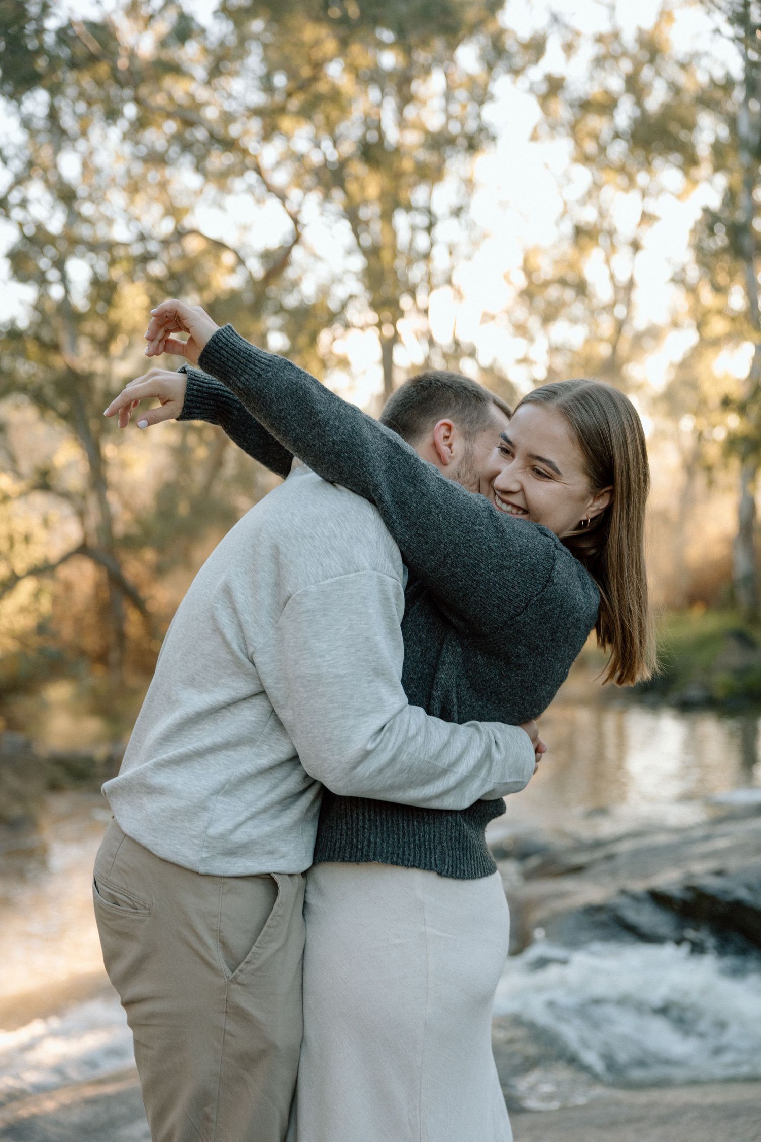 Couple embracing and laughing outdoors near a river, surrounded by autumn trees in warm golden light.