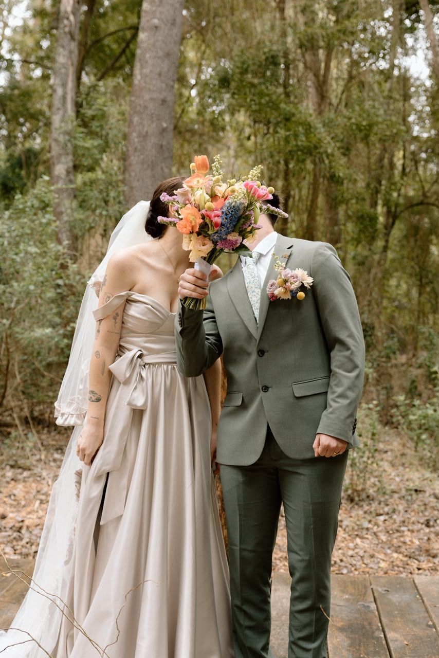 Bride in lavender gown and groom in sage suit playfully hiding faces behind colorful wildflower bouquet in forest.