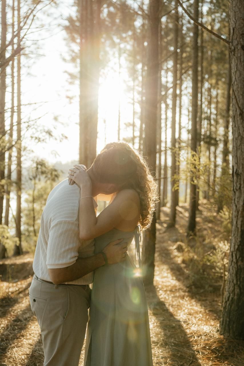 Couple sharing a romantic kiss in a sunlit forest with golden light streaming through tall pine trees.
