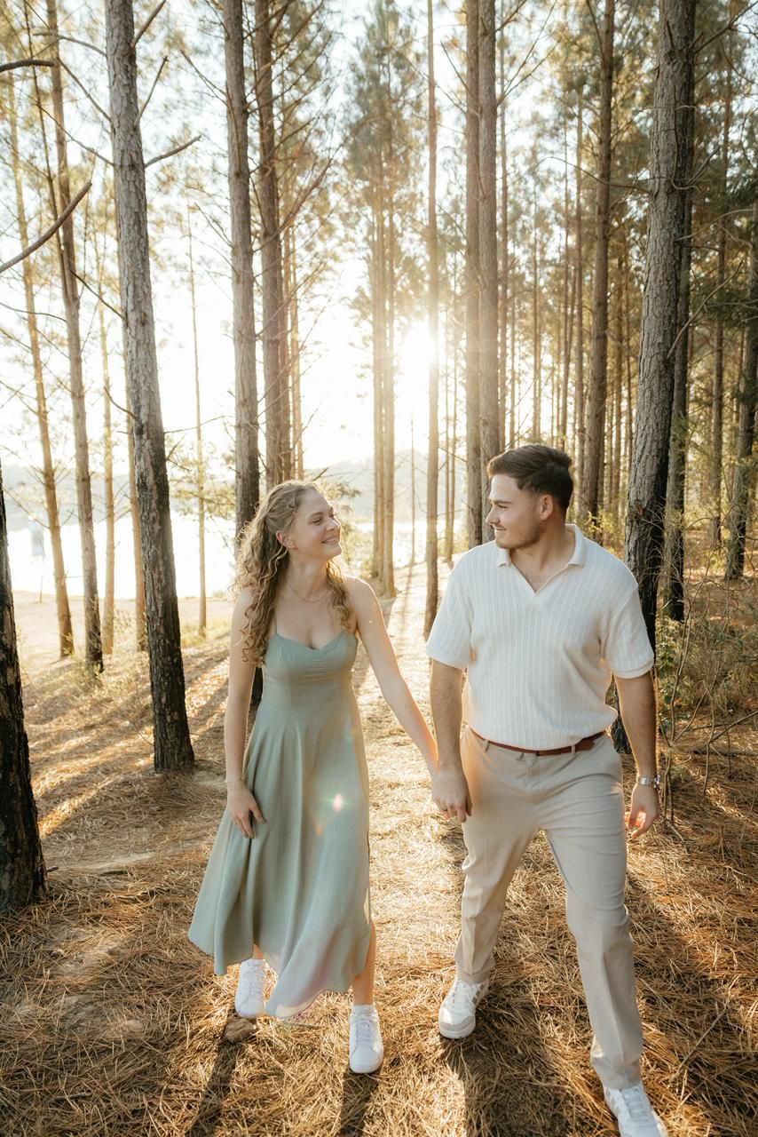Couple holding hands walking through sunlit pine forest in sage dress and casual attire.