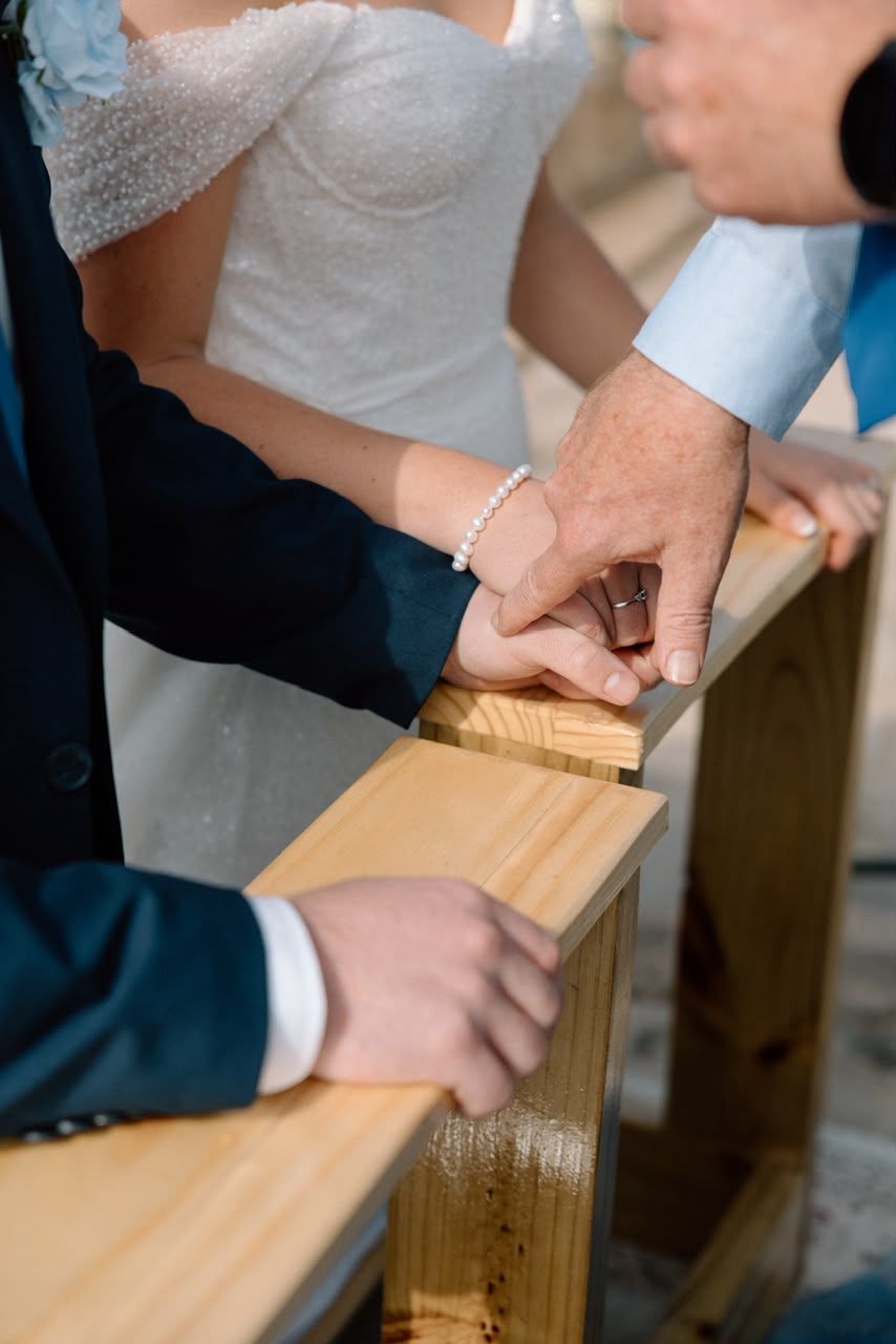 Bride and groom holding hands at a wooden church pew during a wedding ceremony.