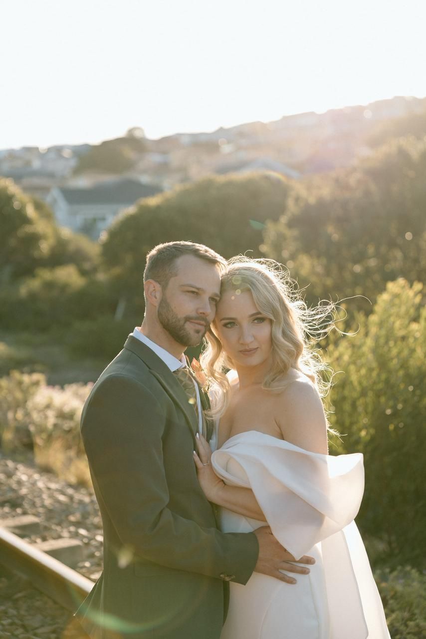 Couple embraces in golden sunset light, bride in off-shoulder white gown and groom in sage green suit.