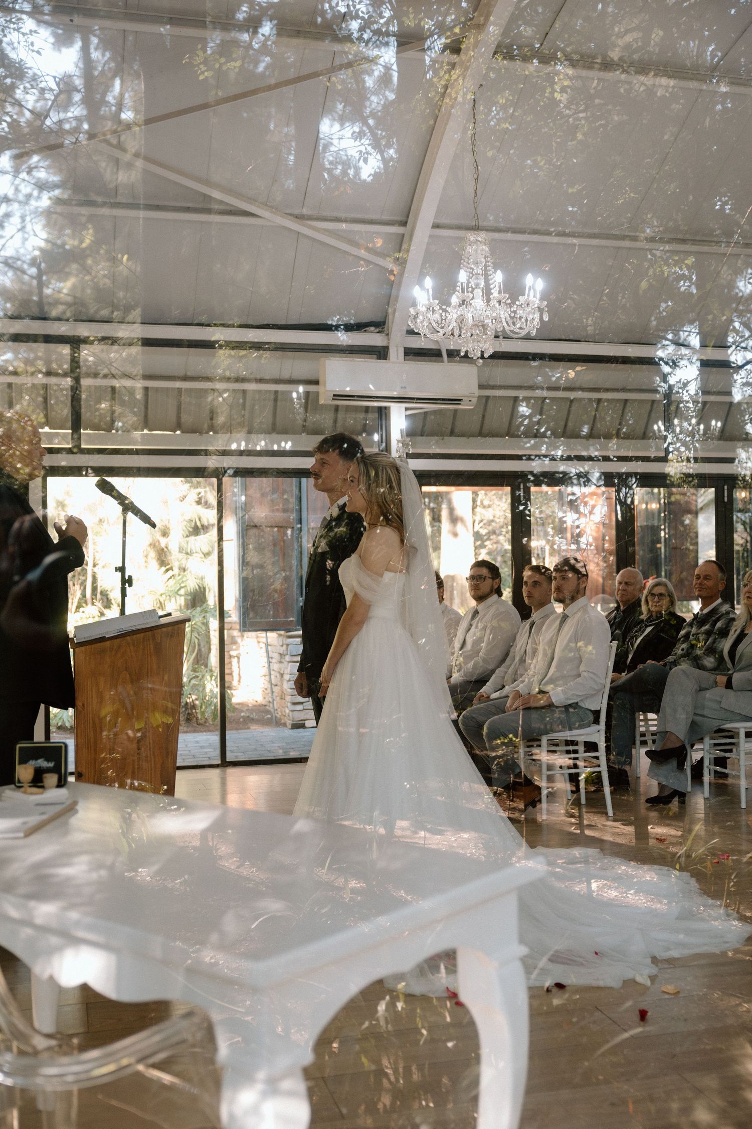Bride in white gown stands with groom during outdoor wedding ceremony under clear tent with chandelier and seated guests.
