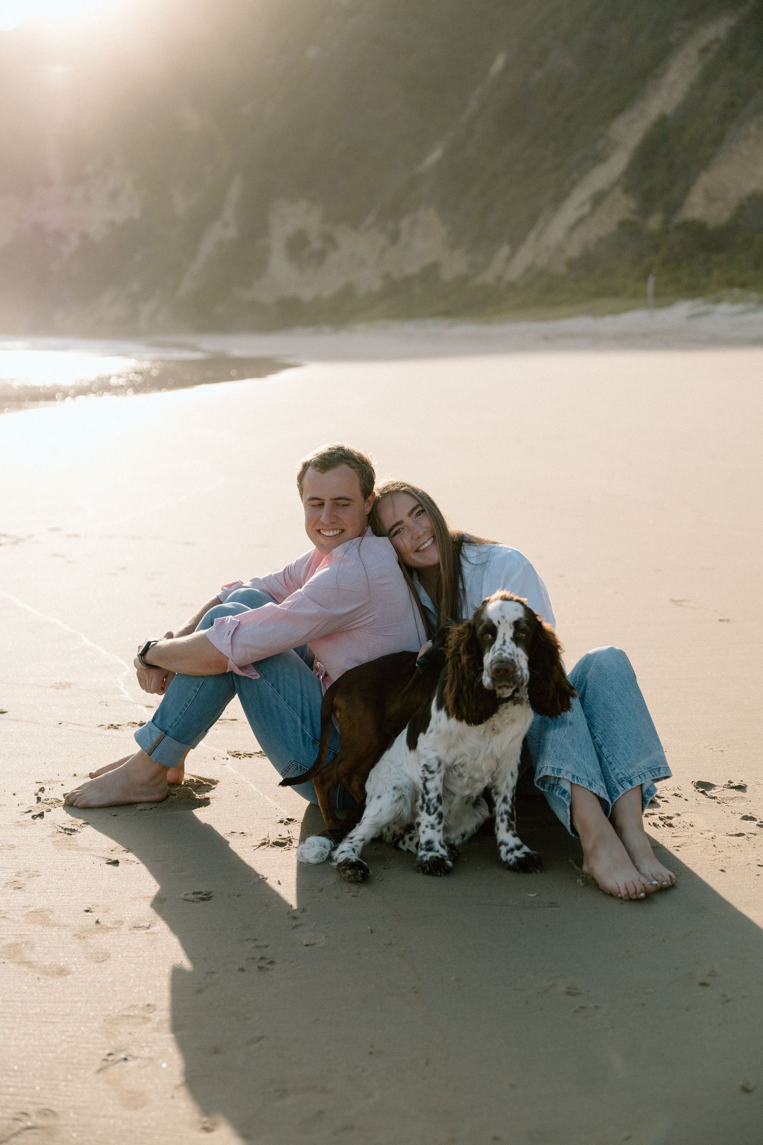 Happy couple sitting on beach sand with their Springer Spaniel dog, leaning together at sunset.
