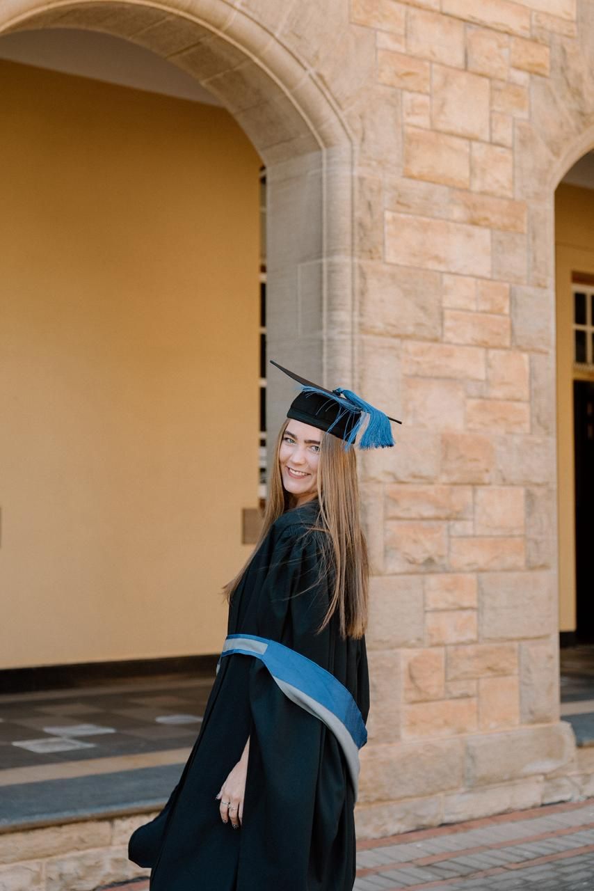 Smiling female graduate in black cap and gown turns to camera beside stone arch.