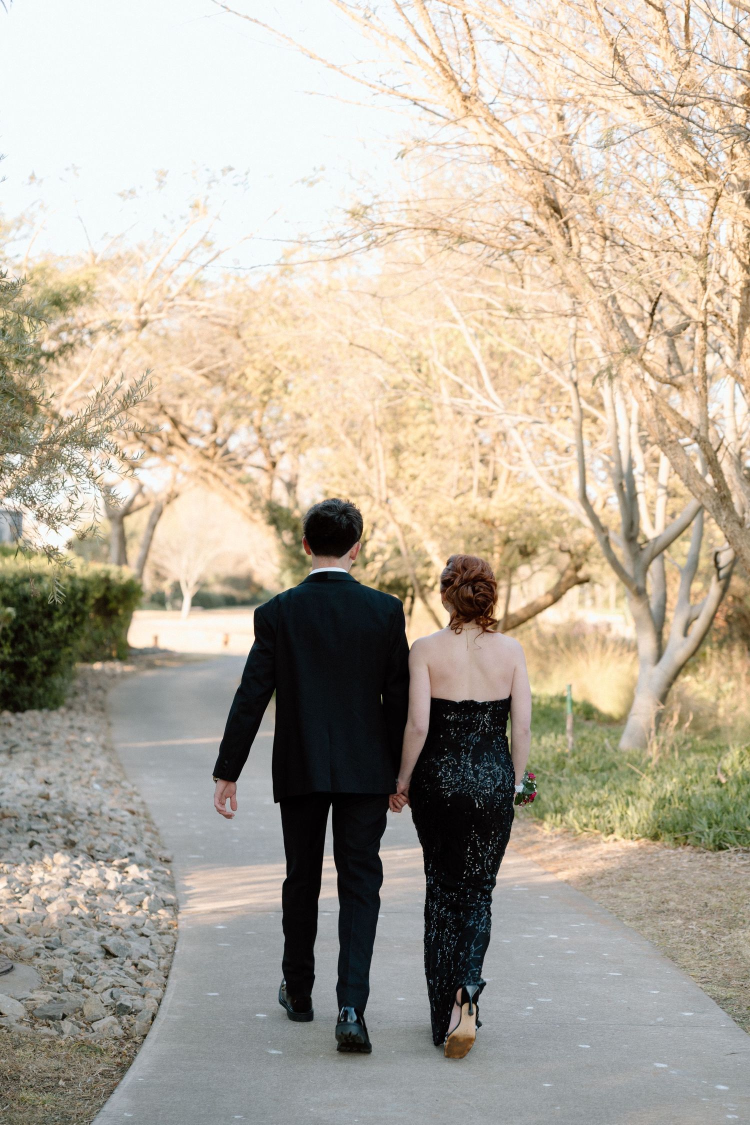 Couple in formal black attire holding hands while walking down a tree-lined path during golden hour.