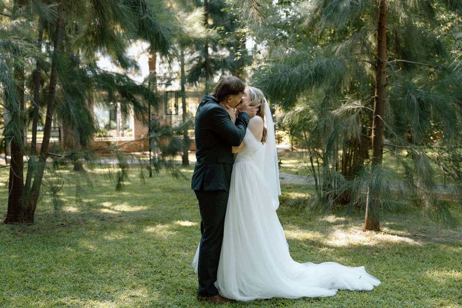 Bride and groom share a romantic kiss surrounded by lush green trees in an outdoor wedding ceremony.
