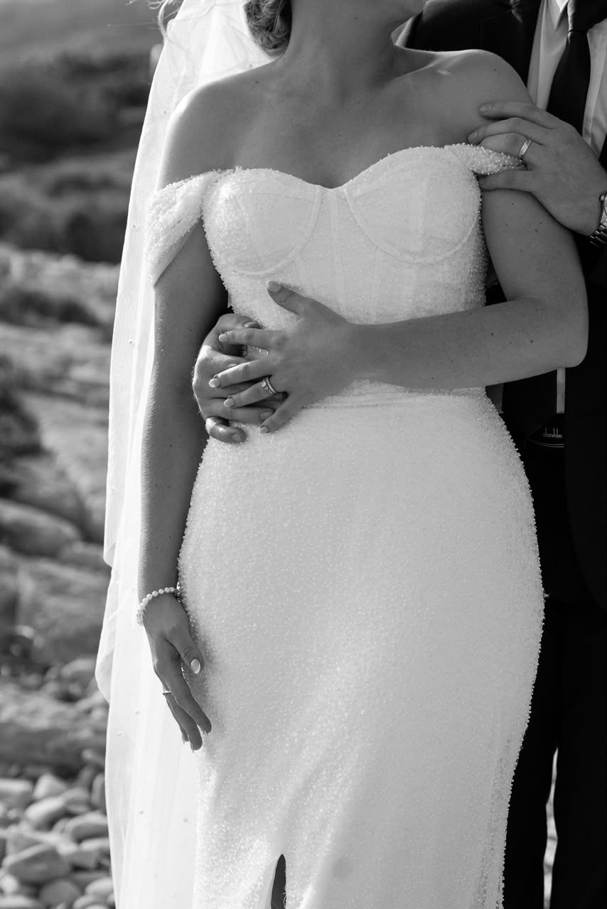 Groom embracing bride in strapless pearl wedding dress with veil, showing wedding rings, in black and white.
