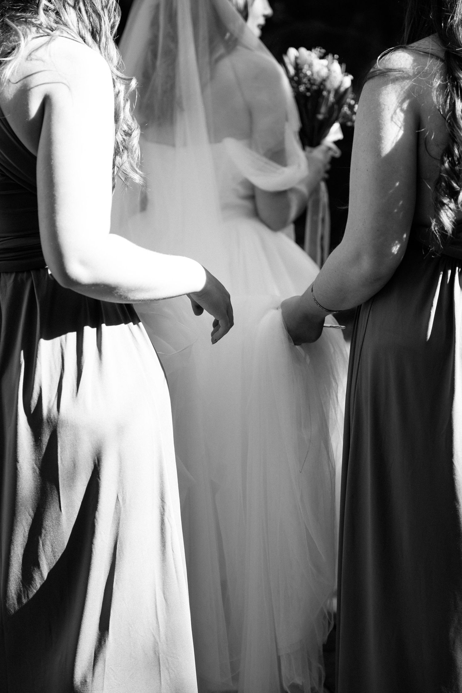 Black and white photo of a bride with bridesmaids in long gowns holding hands at an outdoor wedding ceremony.