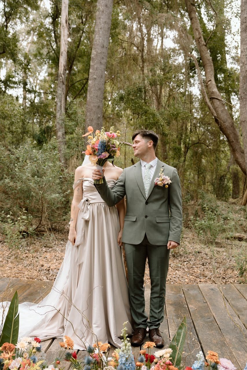 Bride in taupe gown holds colorful bouquet beside groom in green suit on woodland deck.