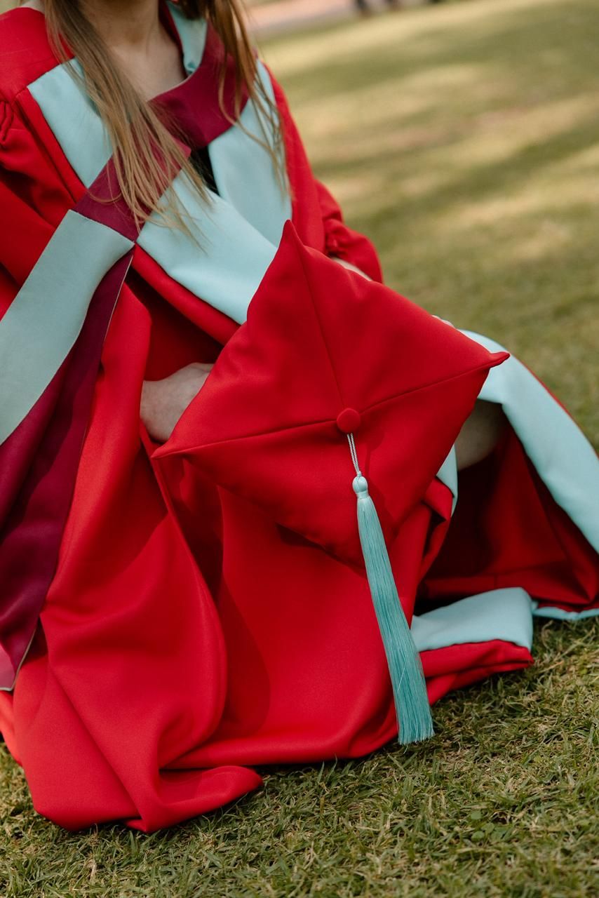 Close-up of red doctoral graduation gown and cap with teal tassel resting on grass.