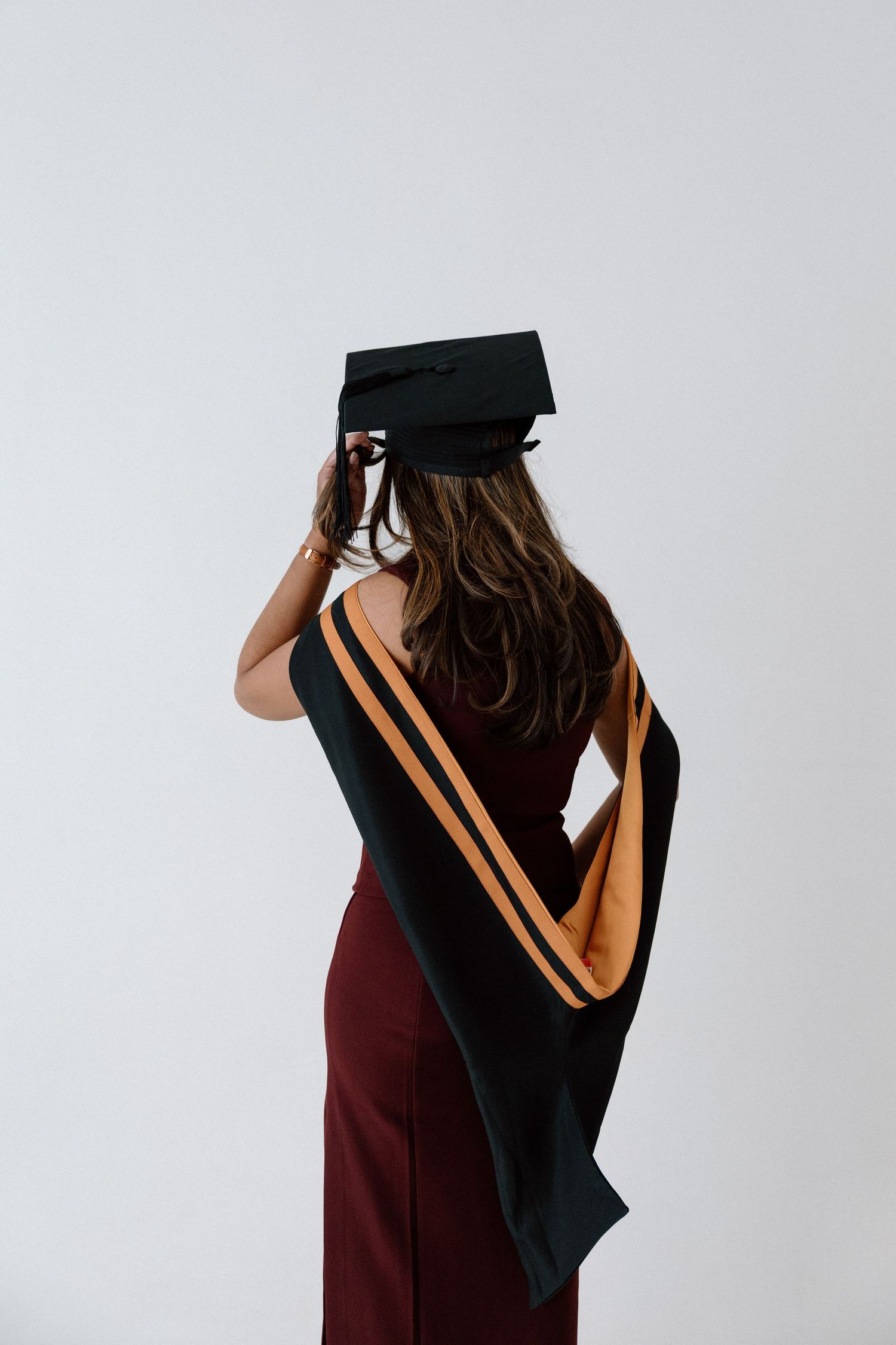 Woman in burgundy dress wearing black graduation cap and hood with gold trim, posed from behind on white background.