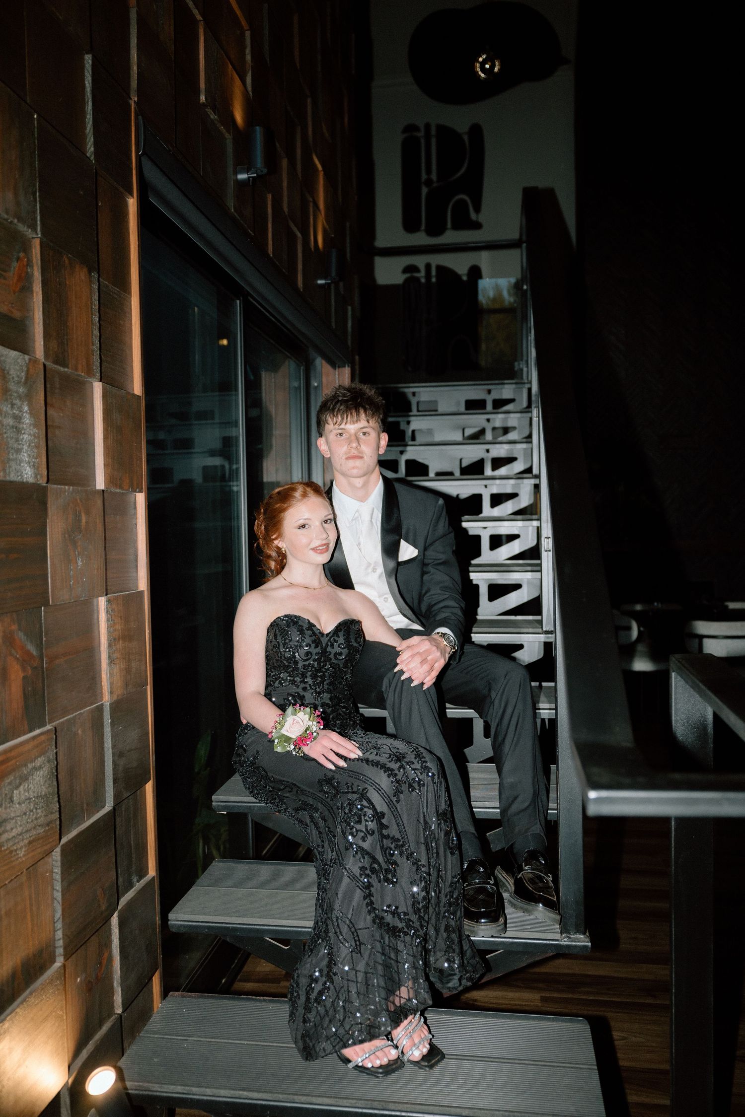 A young couple in formal attire poses on stairs, wearing black gown and suit for prom night.