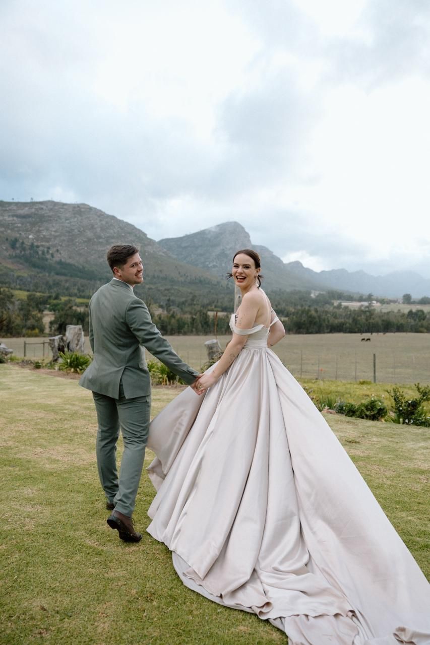 Smiling bride in elegant ball gown holds grooms hand walking through green countryside with mountain views.