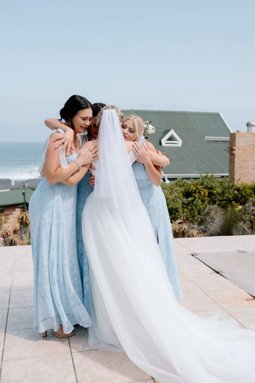 Bride in white gown and veil sharing group hug with two bridesmaids in blue dresses by ocean view.