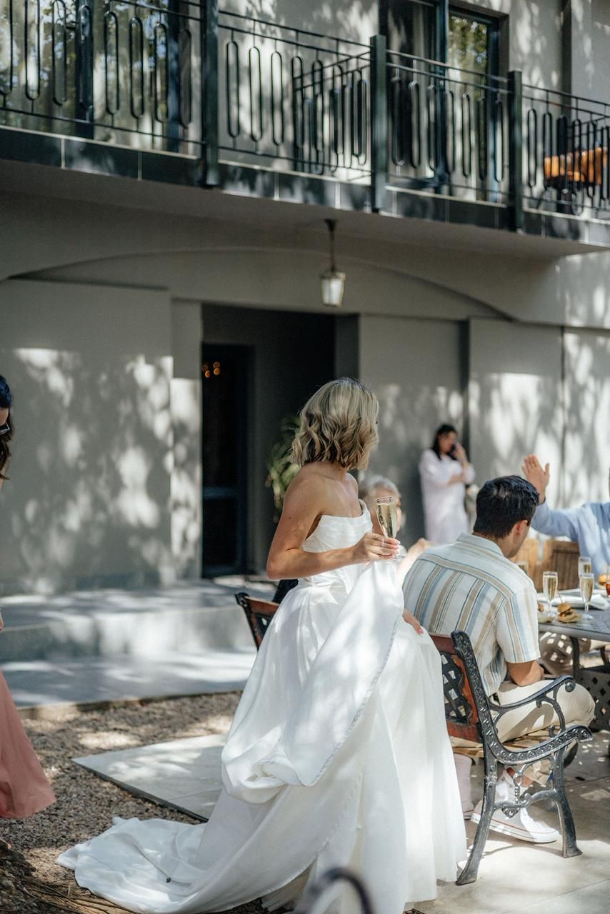 Bride in white strapless gown sits outdoors at reception table holding champagne glass in sunlight.