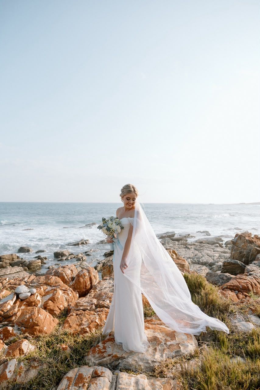Bride in flowing white off-shoulder gown with veil stands on rocky coastal shoreline by ocean.