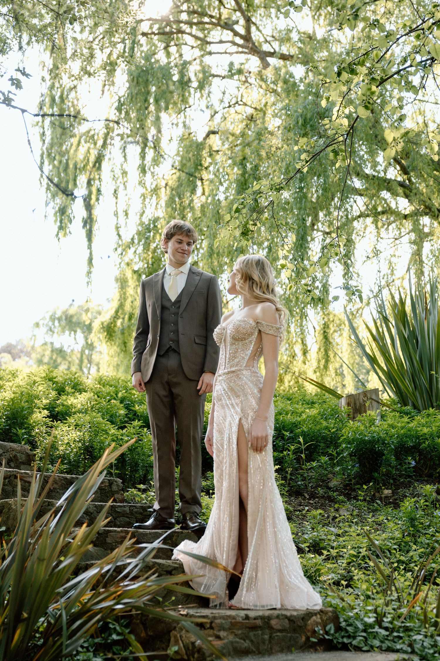 Bride in lace gown and groom in gray suit stand together in lush garden under weeping willow tree.